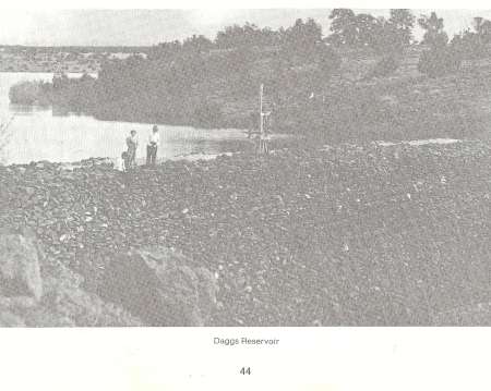 Three people stand near a body of water at Daggs Reservoir, with rocky terrain in the foreground and trees in the background.