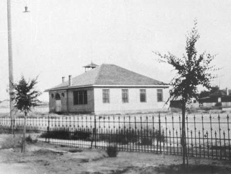 Black and white photo of a small, single-story schoolhouse with a hipped roof, bell tower, fenced yard, and sparse trees in front.