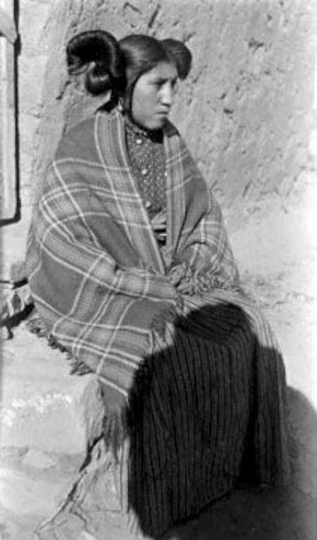 A woman with traditional Hopi hairstyle sits outdoors, wrapped in a plaid blanket and wearing a patterned dress, against a rough textured wall.