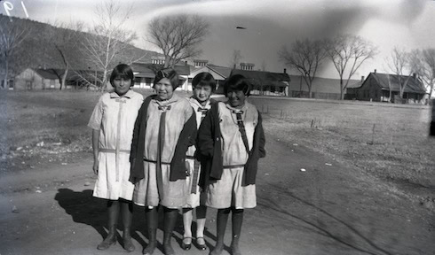 Four girls stand side by side outdoors, wearing similar dresses and jackets, with buildings and bare trees in the background.
