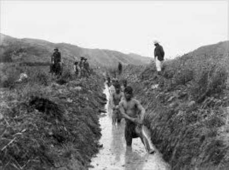 Several men, some shirtless, work digging and clearing a narrow trench or canal in a rural landscape, while others stand observing in the background.