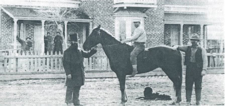 Three men pose with a horse in front of a brick building; one man is seated on the horse, while the other two stand beside it. Several people are visible near the building in the background.