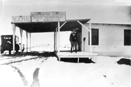 Black and white photo of a man standing on the porch of a Quarantine Station building, with snow on the ground and an old car parked nearby.