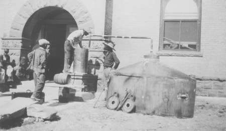 Three men stand beside a large metal still and connected tank outside a stone building with an arched doorway and a window.