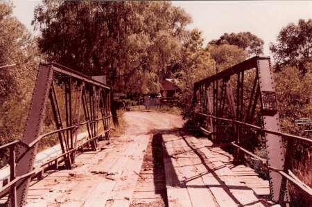 A weathered steel truss bridge with a wooden plank deck crosses over a small creek, surrounded by trees and foliage.