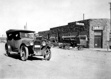 Black-and-white photo of early 20th-century cars parked on a dirt road in front of a stone theater and a building labeled "Merchants.