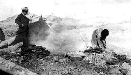 A man stands while a woman crouches and works with stones near a bowl on rocky ground in a barren outdoor setting.