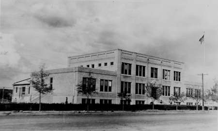 Black-and-white photo of a two-story brick school building with an American flag on a pole, trees lining the street, and overcast skies in the background.