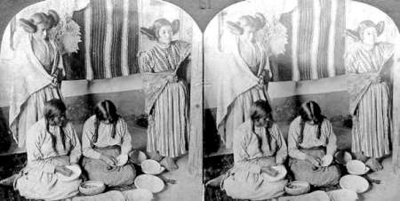 Four women, two standing and two seated, prepare food with bowls and baskets in a room decorated with patterned textiles.