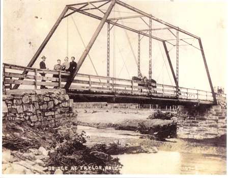 Black-and-white photo of a truss bridge with four people and a horse-drawn carriage on it, spanning a small creek with rocky banks.