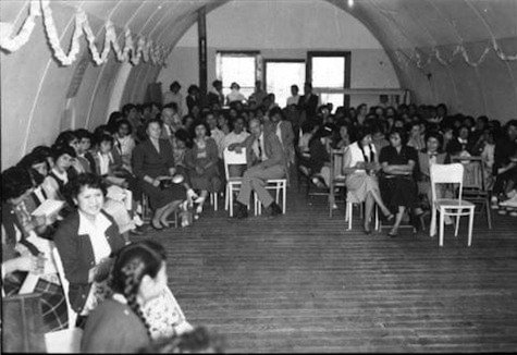 A large group of people, including children and adults, sit on chairs in rows inside a decorated hall with arched ceilings, facing the camera.