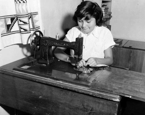 A young girl operates a vintage sewing machine at a wooden table in a room, concentrating on her work.
