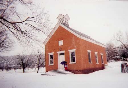 A small brick building with a steeple stands in a snowy landscape; a person with a blue umbrella is near the entrance steps.