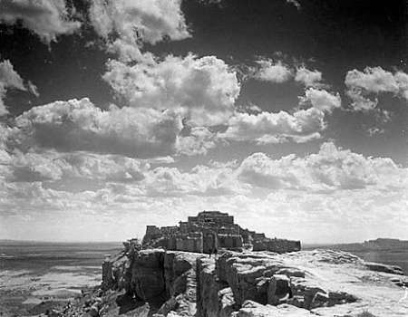 A stone pueblo sits atop a rocky mesa under a sky filled with scattered clouds, overlooking a vast and open landscape.