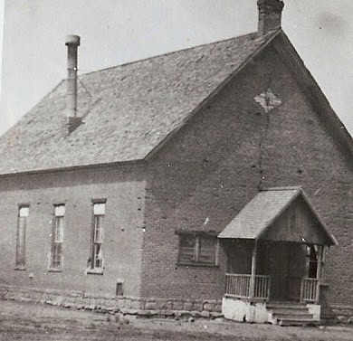 Black-and-white photo of a small, old brick building with a pitched roof, three tall windows, a chimney, and a covered front porch.