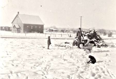 A man, two children, and a dog stand in a snow-covered yard near a pile of wood, with a house and outbuildings in the background.