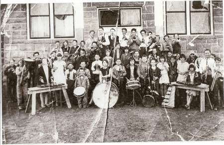 A large group of children and adults pose outdoors with various musical instruments in front of a brick building with two windows.