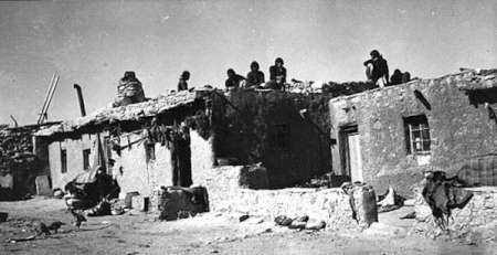 Several people sit on the flat roof of an adobe building in a sunlit, arid setting. The structure shows signs of wear, with bags and containers nearby on the ground.