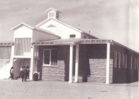 A black-and-white photo of a single-story school building with a covered porch; a few children stand near the entrance.