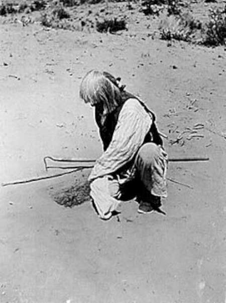 A person kneels on sandy ground, using a stick to dig or examine the earth, with sparse vegetation visible in the background.