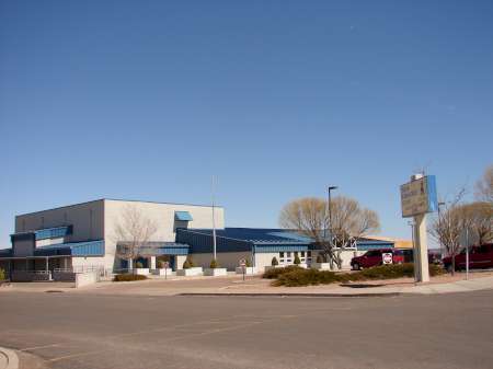 A modern, single-story building with blue trim and awnings sits beneath a clear sky, with a parking lot and a sign in front.