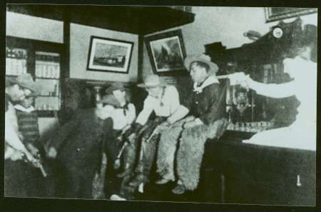 Historic black-and-white photo showing four men with hats and guns sitting in a room while a woman points a pistol at them. The setting appears to be the American Old West.