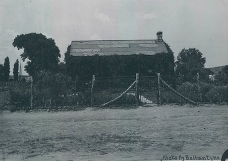Black and white photo of a small house with a pitched roof, covered in vines, behind a wooden fence and gate, surrounded by trees and shrubs.