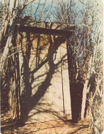 A weathered wooden outhouse stands surrounded by leafless branches in a natural outdoor setting.