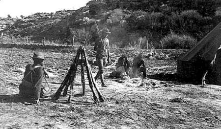 Several soldiers are gathered at a campsite with rifles stacked in a teepee formation; a tent and rocky terrain are visible in the background.