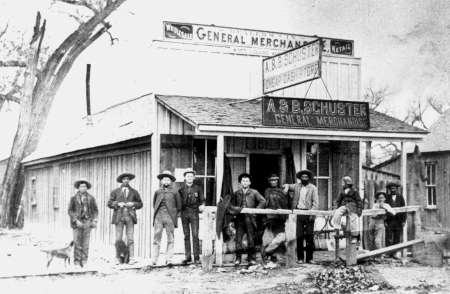 Black and white photo of people standing in front of a wooden general merchandise store with signs reading "A. & B. Schuster General Merchandising.