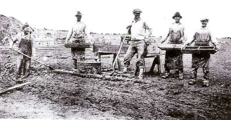 Five men stand outdoors with mining equipment and soil, appearing to sift or pan for minerals, likely during an early 20th-century mining operation.