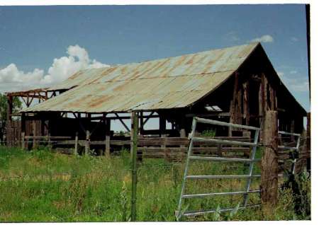 An old, weathered barn with a rusty metal roof sits in a grassy field; a metal gate leans against a wooden fence in the foreground.