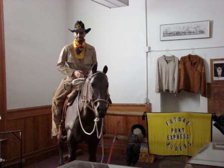 A person dressed in cowboy attire sits on a horse indoors next to a yellow "Future Pony Express Riders" sign and cowboy-themed clothing displays.