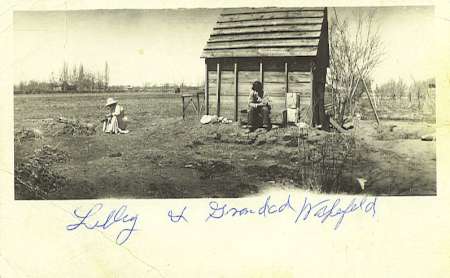 Two children sit outside a small wooden shed in a barren field. One child sits on a bench near the shed; the other sits on the ground. Handwritten text is on the photo's lower edge.