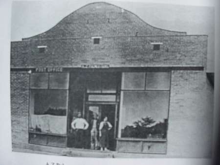 Black and white photo of three people standing in front of a brick building with large windows and a sign that reads "Post Office.