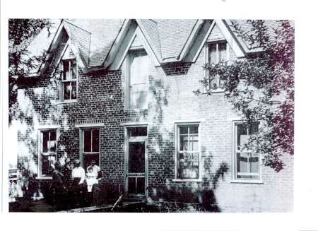 Black and white photo of a brick house with steep gabled roofs; two people are seated near the entrance, partially shaded by tree branches.