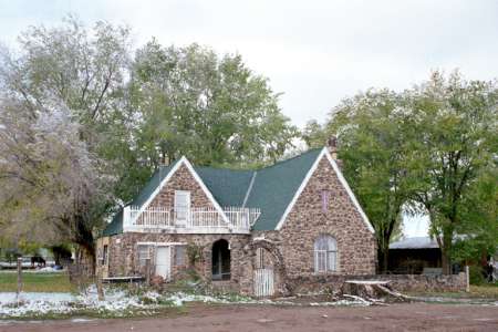 A stone house with a steep green roof, white trim, and a partially fenced front yard, surrounded by trees and patches of snow on the ground.