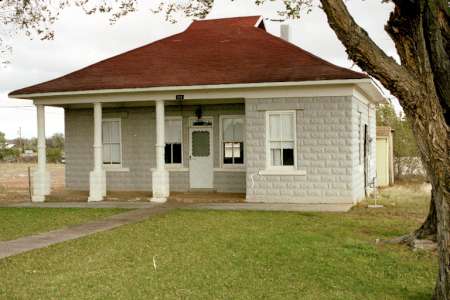 Single-story house with a red hipped roof, gray brick exterior, front porch supported by four columns, and a large tree on the right side of the lawn.
