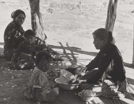 Four people, including two women and two children, sit on the ground outdoors under the shade of trees, with bowls and kitchenware in front of them.