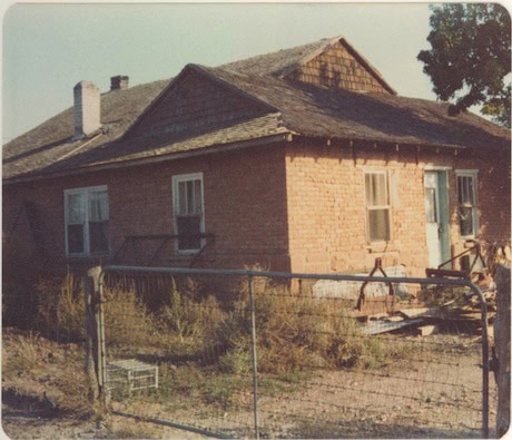 A weathered brick house with a shingled roof, overgrown yard, and metal gate in front. A tree is visible in the background.