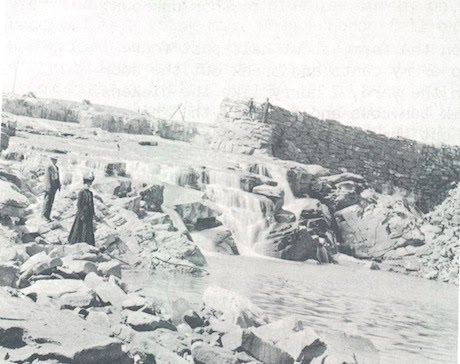 Black and white photo of two people standing near a rocky stream with a small waterfall and a stone wall in the background.