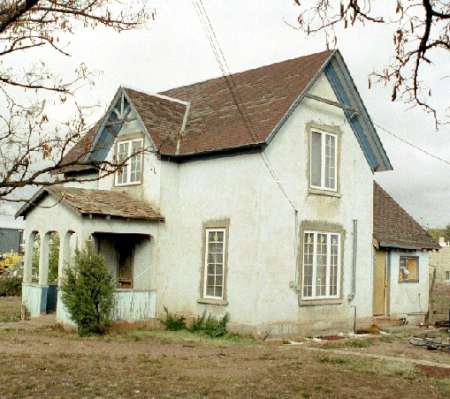 A small, old, two-story house with faded light blue paint, a covered porch, and a sloped roof, surrounded by bare trees and sparse vegetation.