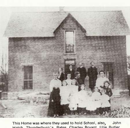 A group of adults and children pose in front of a two-story brick house; the photo is black and white and appears historical.