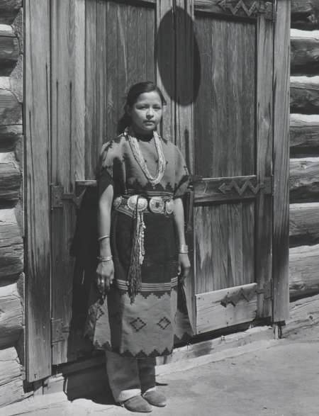 A woman wearing traditional clothing and jewelry stands in front of wooden double doors on a log building.