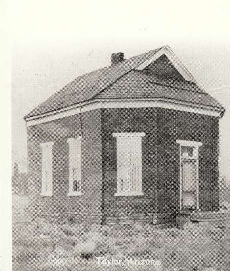 Black and white photo of a small brick building with three tall windows and a single door, standing on rough ground. The text "Taylor, Arizona" is faintly visible at the bottom.