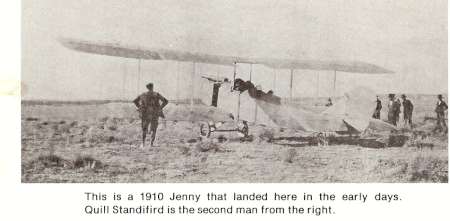 A group of men stand near a 1910 Jenny biplane in an open field; the plane appears to have just landed.
