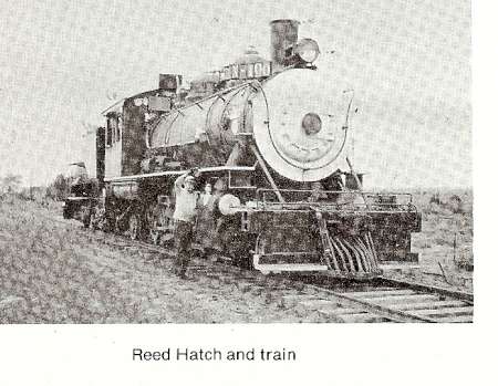 Black-and-white photo of a steam locomotive stopped on railroad tracks with a man standing next to the front of the train.