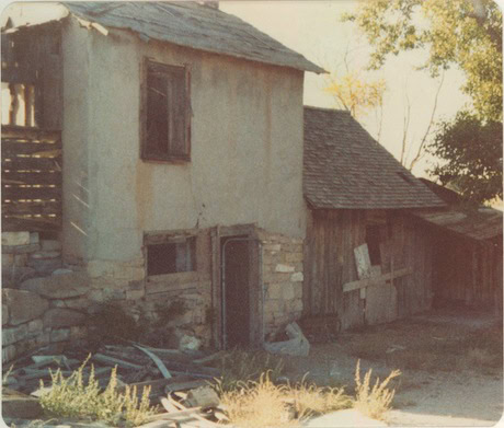Old, weathered two-story building with boarded windows and doors, a partially collapsed roof, and overgrown weeds in the foreground.