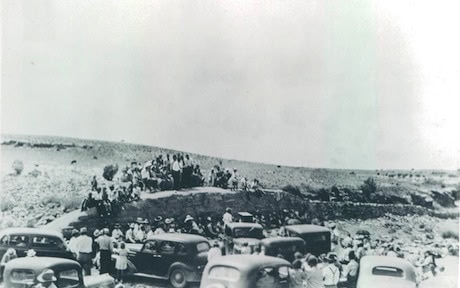 A black-and-white photo of a crowd gathered near parked cars on a barren landscape, with people standing on an elevated area in the background.
