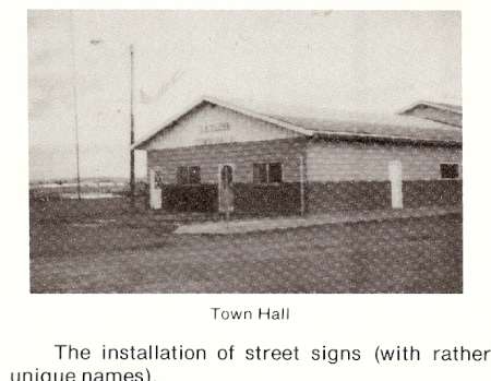 A small, single-story building labeled "Town Hall" stands on a dirt lot with a flagpole nearby.
